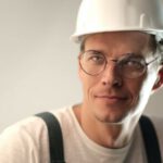 Safety Construction Vest - Content male builder in workwear and hardhat smiling on gray background in studio and looking at camera