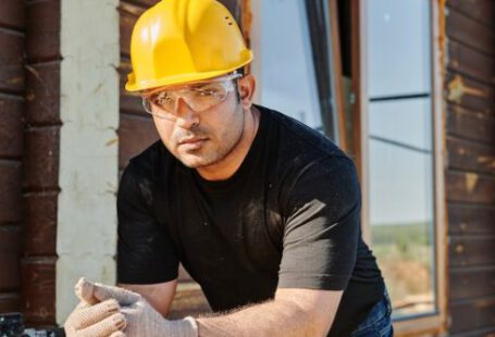 Work Gloves - Man in Black Crew Neck T-shirt Wearing Yellow Hard Hat While Standing by the Wooden Table