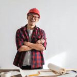 Builder - Smiling casual man in hardhat and glasses holding arms crossed looking at camera while standing at desk with paper draft and stationery
