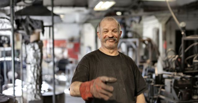 Construction Industry - Cheerful senior white hair craftsman in heavy duty gloves laughing in workshop while looking at camera