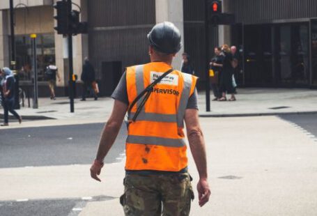Safety Construction Vest - Back view of unrecognizable young male constructor in protective helmet and vest walking on asphalt road in modern city district
