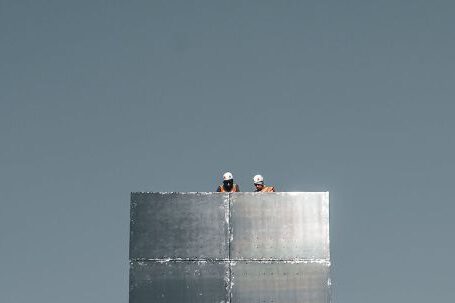 Construction - From below of anonymous distant engineers in helmets working on rooftop of modern skyscraper with glass mirrored walls against cloudless blue sky