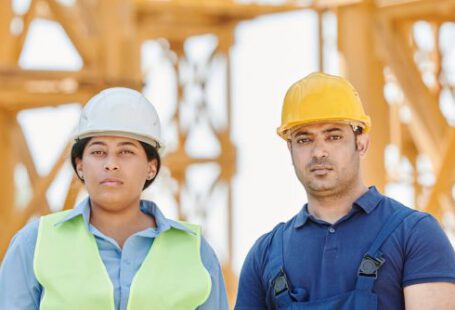 Construction Workers - A Man and a Woman Wearing Hardhats Standing Near a Metal Structure