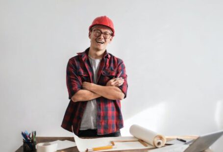 Builder - Smiling casual man in hardhat and glasses holding arms crossed looking at camera while standing at desk with paper draft and stationery