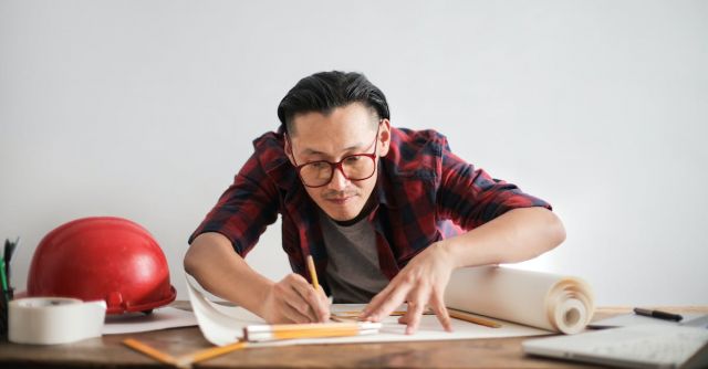 Builder - Inspired young male engineer in glasses drawing draft on paper bending over table against white wall